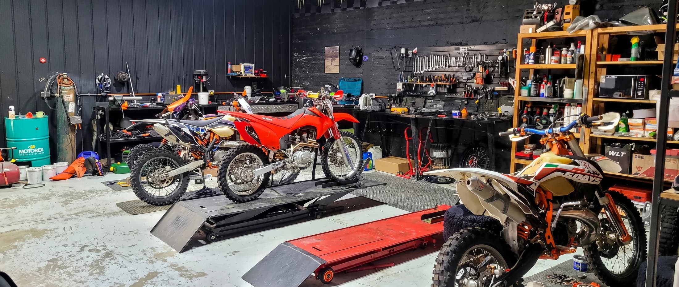 Interior del taller de Proride con dos motos de enduro sobre el elevador hidráulico, el muro de herramientas ordenado al fondo y una KTM en mantención en primer plano