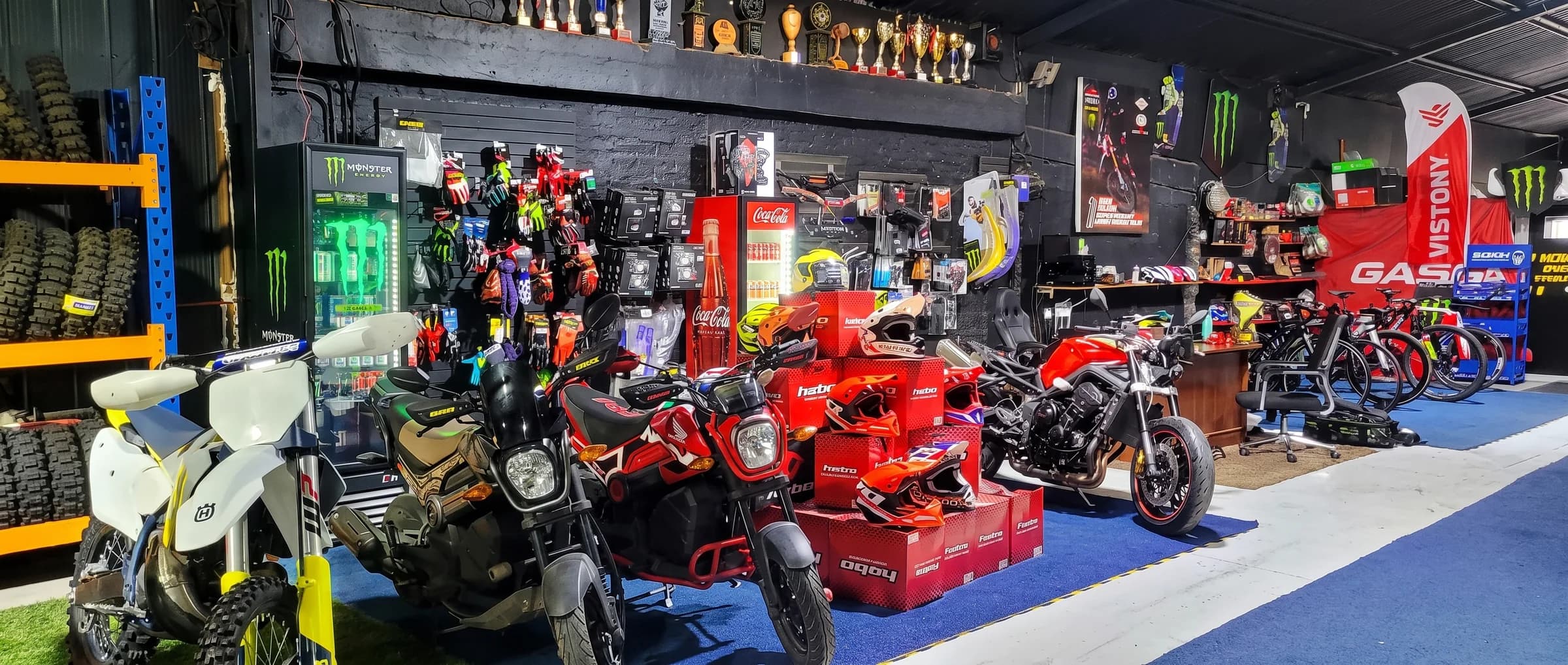Vista panorámica del local de Proride: motos de enduro, calle y una naked en exhibición, muro de guantes y accesorios, cascos apilados y la repisa de trofeos de competencia sobre la pared negra